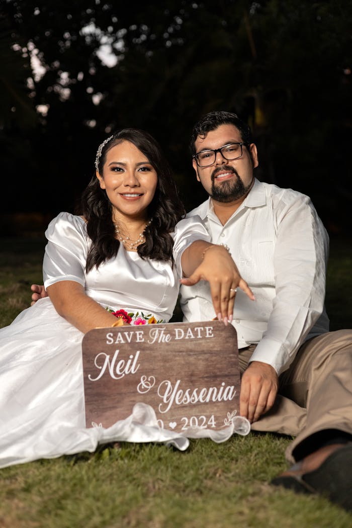 Happy couple posing with a 'Save the Date' sign in an outdoor setting during the day.